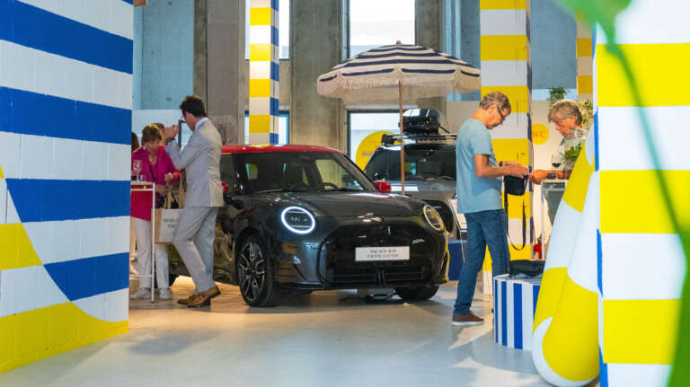 Voiture MINI noire exposée à l’intérieur, entourée de colonnes rayées et d’éléments estivaux.